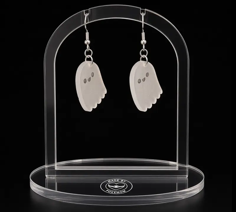 A pair of pale laser-cut ghost earrings with tiny faces, hanging from silver-tone hooks on a clear acrylic stand.