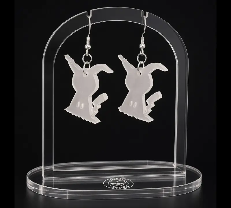 A pair of pale laser-cut Mimikyu-inspired earrings in a disguise shape, hanging from silver-tone hooks on a clear acrylic stand.