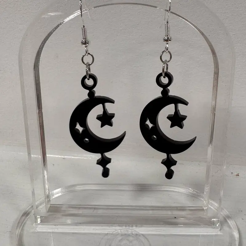 A pair of matte black crescent and falling-star acrylic earrings on silver-tone hooks, displayed on a card.
