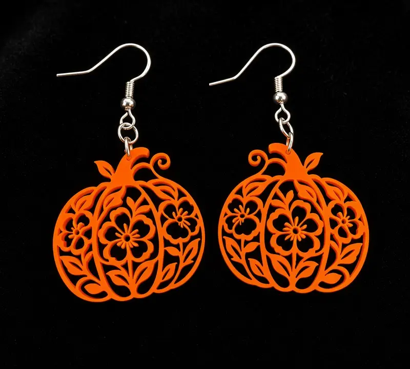 A pair of bright orange laser-cut pumpkin earrings with a lace-like filigree pattern, on silver-tone hooks against a black background.